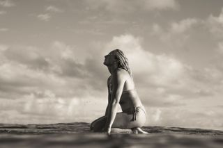 A black and white atmospheric image of pro-surfer Izzi Gomez sitting on her board in the sea. She is kneeling on the surfboard, which is also called kneeboarding, and she is looking up towards the sky.