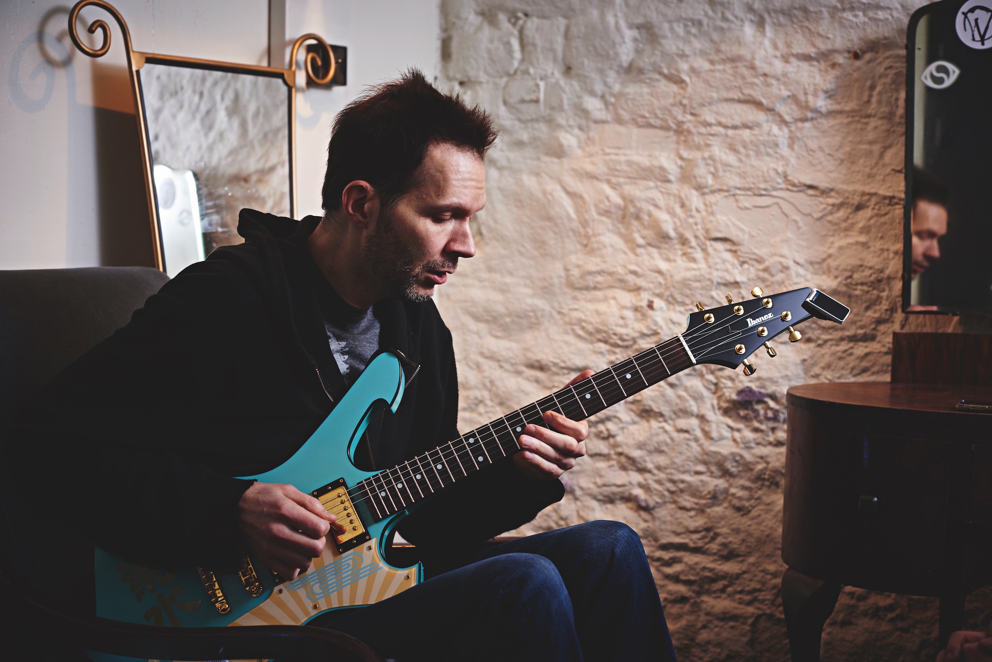 Paul Gilbert, photographed seated with a light blue-finished example of his signature Ibanez Fireman guitar