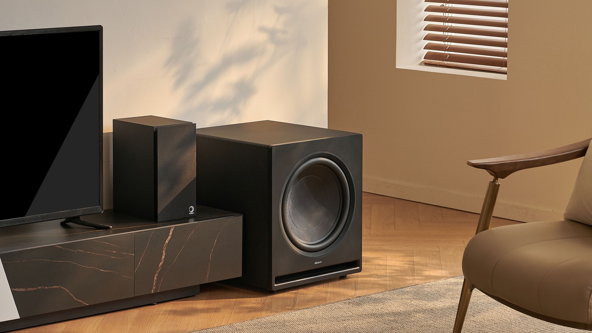 A lifestyle photo of the Elipson Premium SUB next to a TV and speaker in a neutrally decorated room