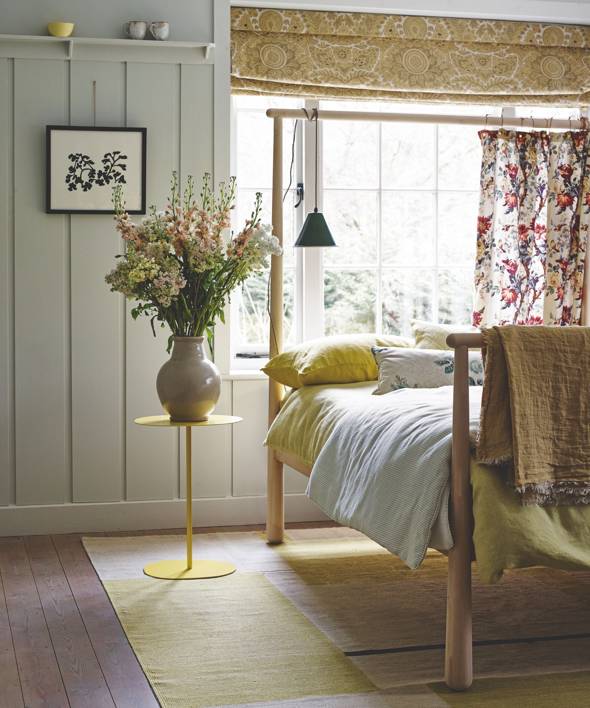 A bright bedroom with large windows, four poster bed, yellow bedding, yellow bedside table with large vase of flowers, yellow rug, and dark wood flooring. The walls are pale green panelled.