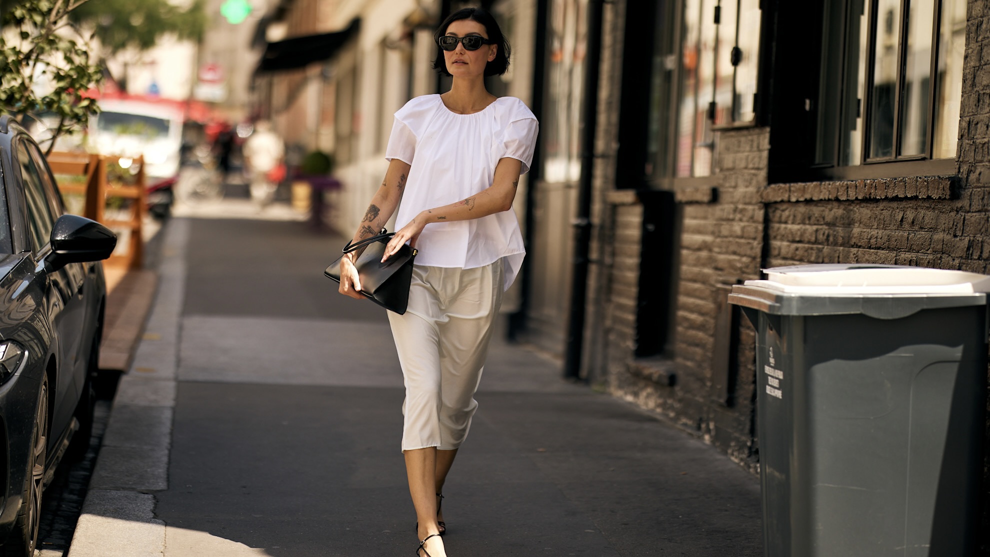 Chic Parisian woman wearing a white top and skirt with black strappy sandals, black bag and black sunglasses
