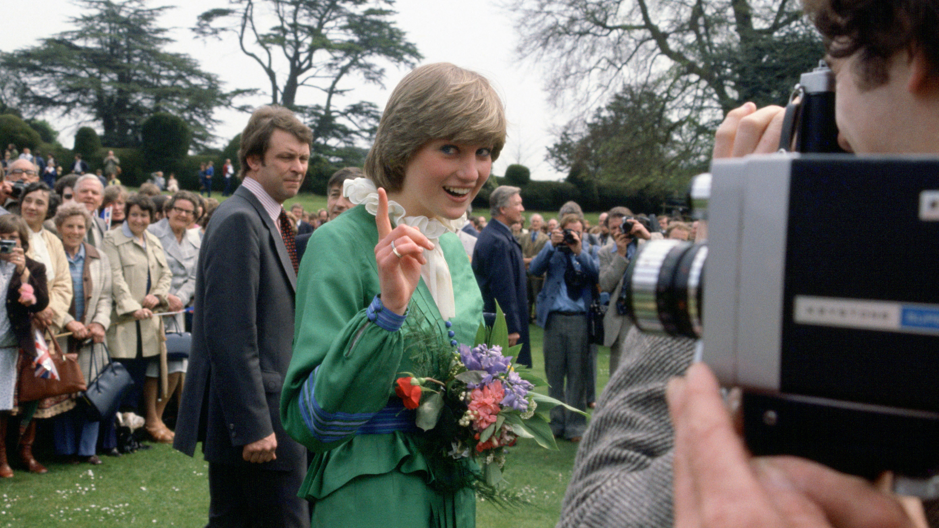 Princess Diana wearing a green dress waving her finger at the camera as a man takes a video of her