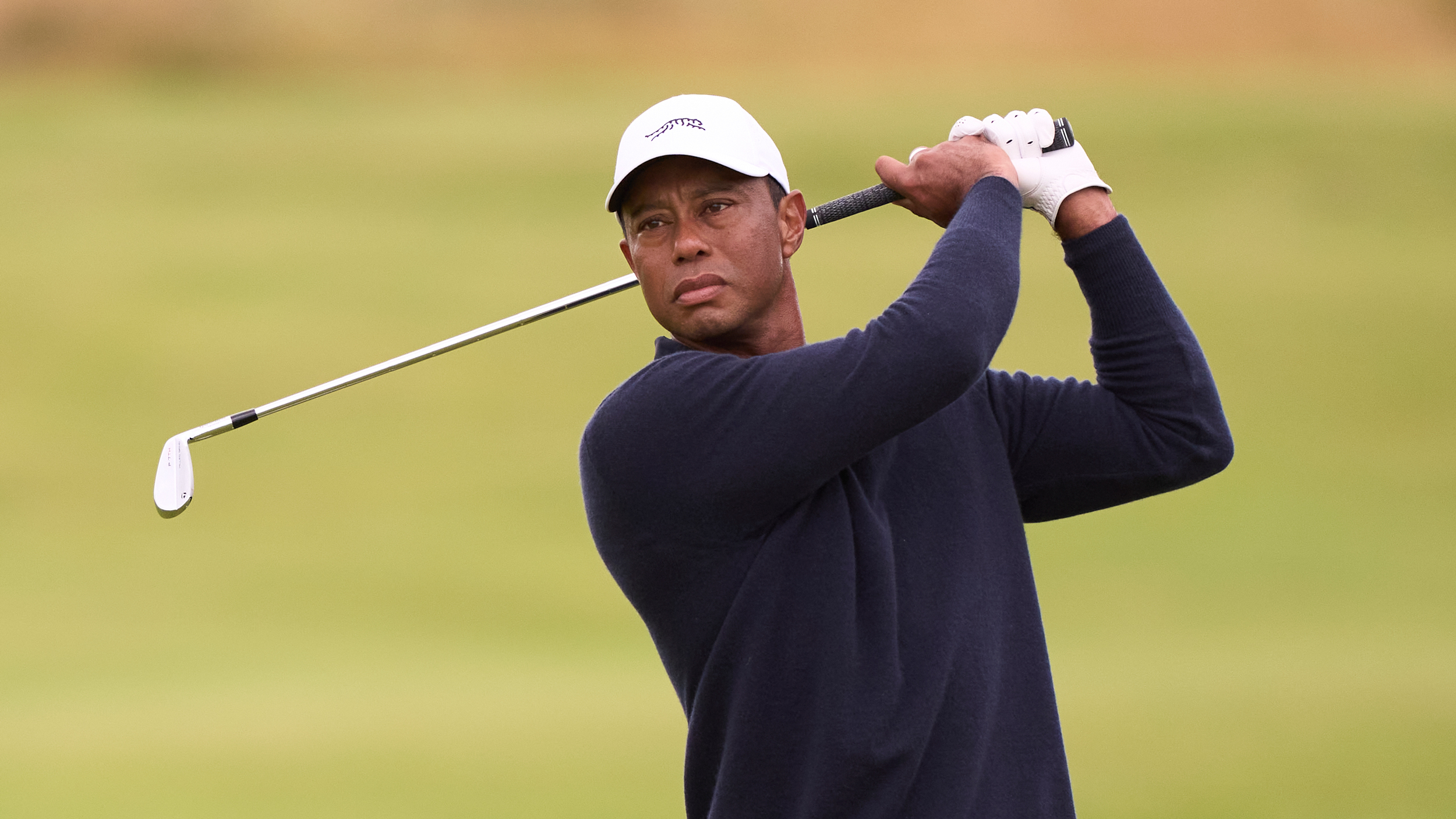 Tiger Woods holds his finish on an iron shot while wearing a navy sweater and white cap