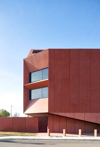 Daytime image of the new contemporary Art Centre, red exterior, road, surrounding trees, red stone wall, small posts in a line on the pathway, blue cloudy sky