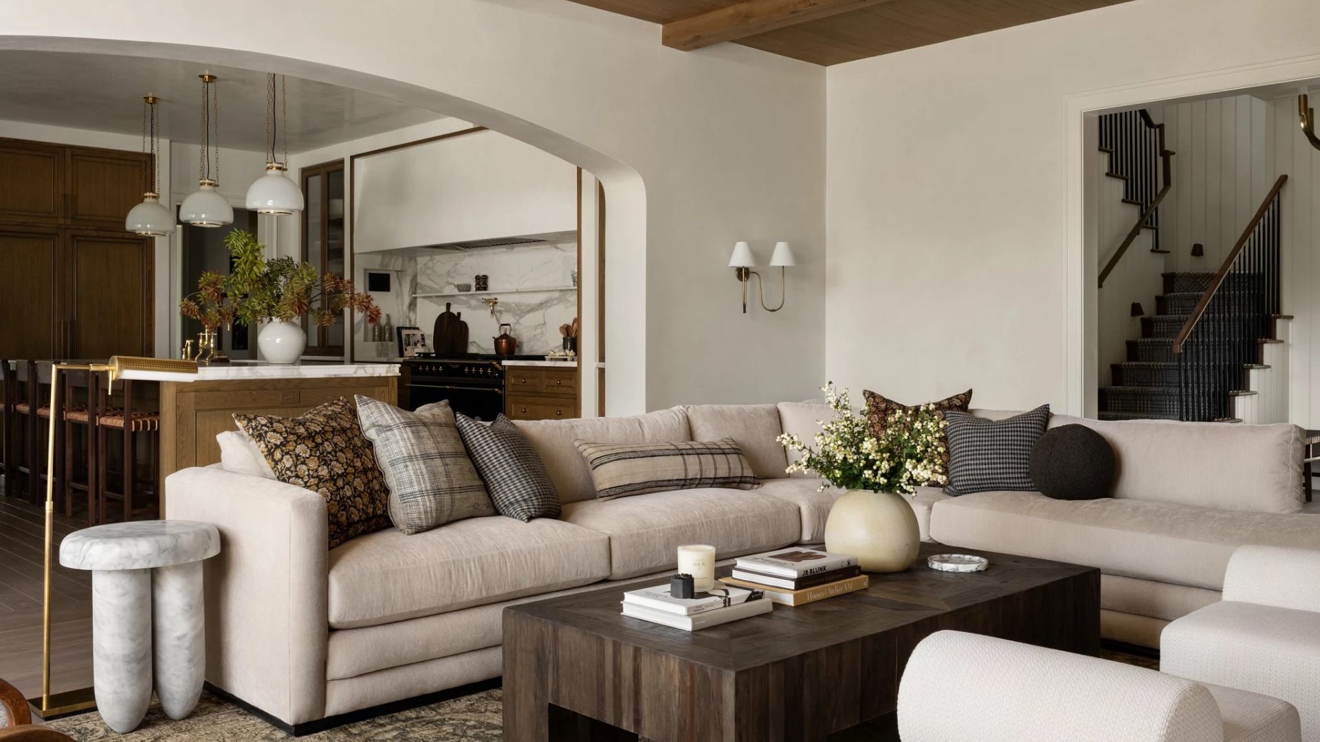 A bright white living room with vaulted beams on the ceiling, wall sconces, a beige sectional with patterned windows, a dark wood coffee table, a marble stool and a staircase