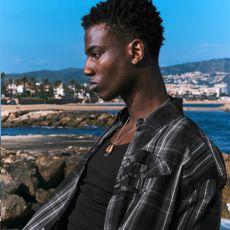 A side profile photo of a male model wearing a black tartan open shirt and a black vest top at the coast