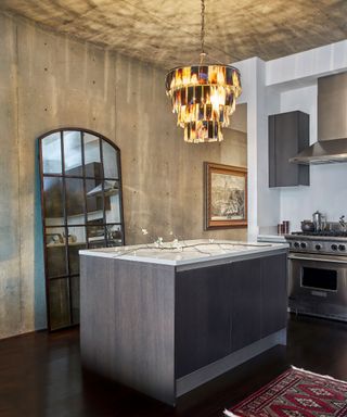 Modern kitchen with concrete style wall and ceiling, dark wood island, dark wood paneled mirror design leaning on wall, feathered chandelier above island