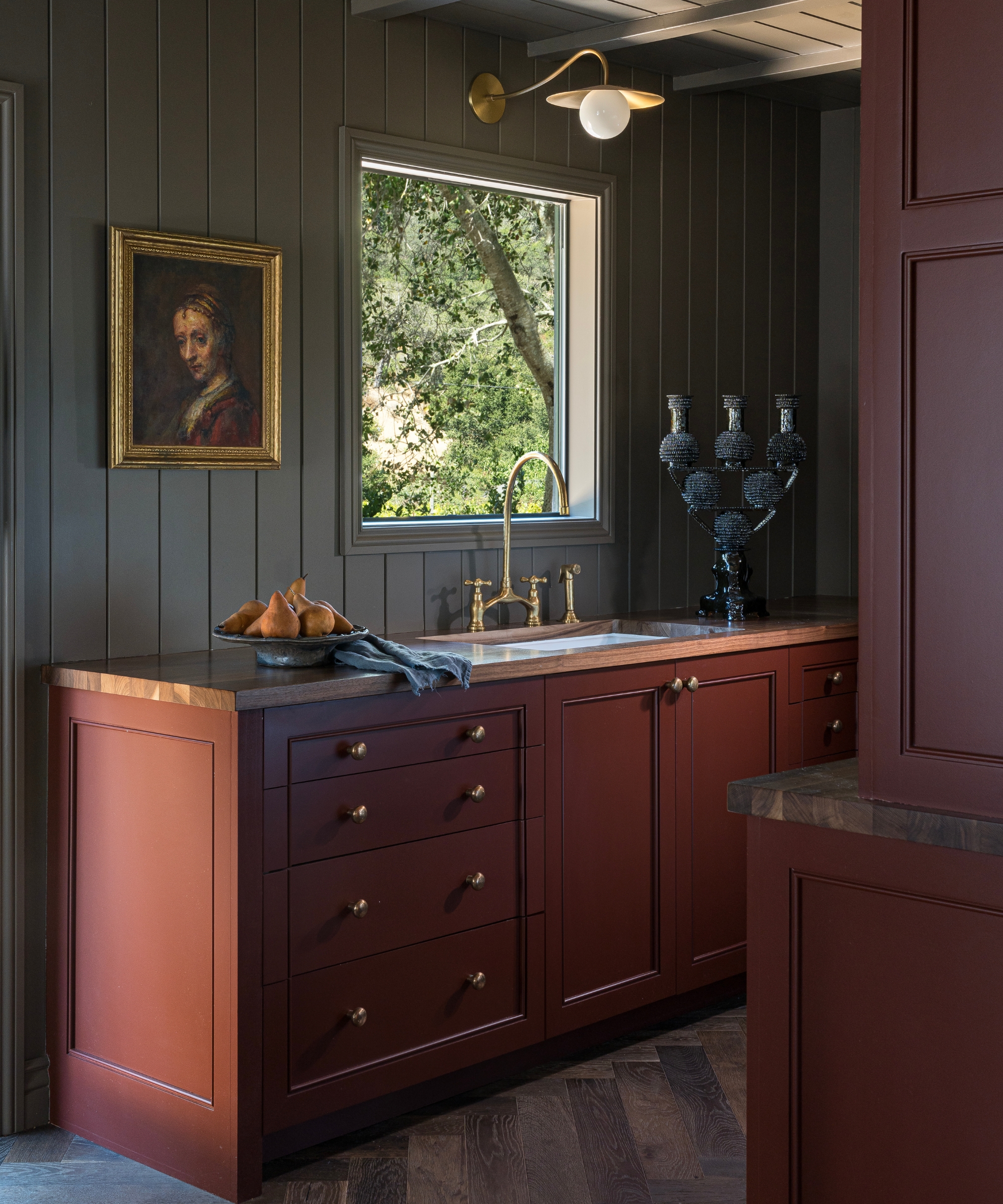 A small cottage kitchen with dark green paneled walls, wooden countertops, and rich red cabinetry with a unique molding design