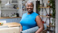 A woman stands in a kitchen with a yoga mat rolled under her arm. She is wearing a sports top and smiling at the camera. Behind her we see modern white brick tiling, open shelving, and green leafy plants in the kitchen.