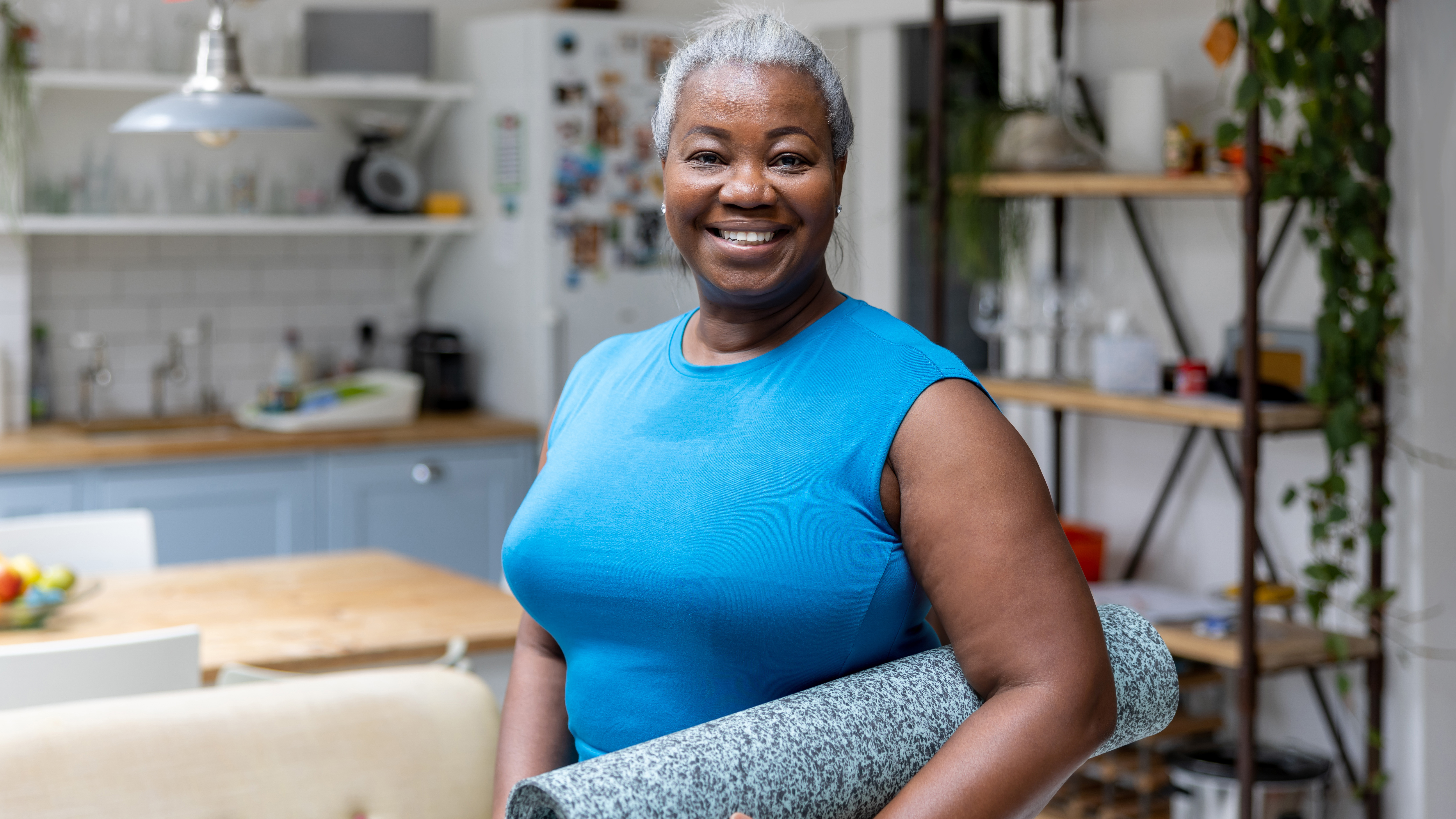 A woman stands in a kitchen with a yoga mat rolled under her arm. She is wearing a sports top and smiling at the camera. Behind her we see modern white brick tiling, open shelving, and green leafy plants in the kitchen. 