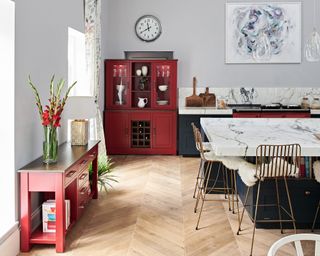 A red cabinet and console table in a kitchen with marbled island, brass-colored bar stools and Herringbone-style wooden flooring