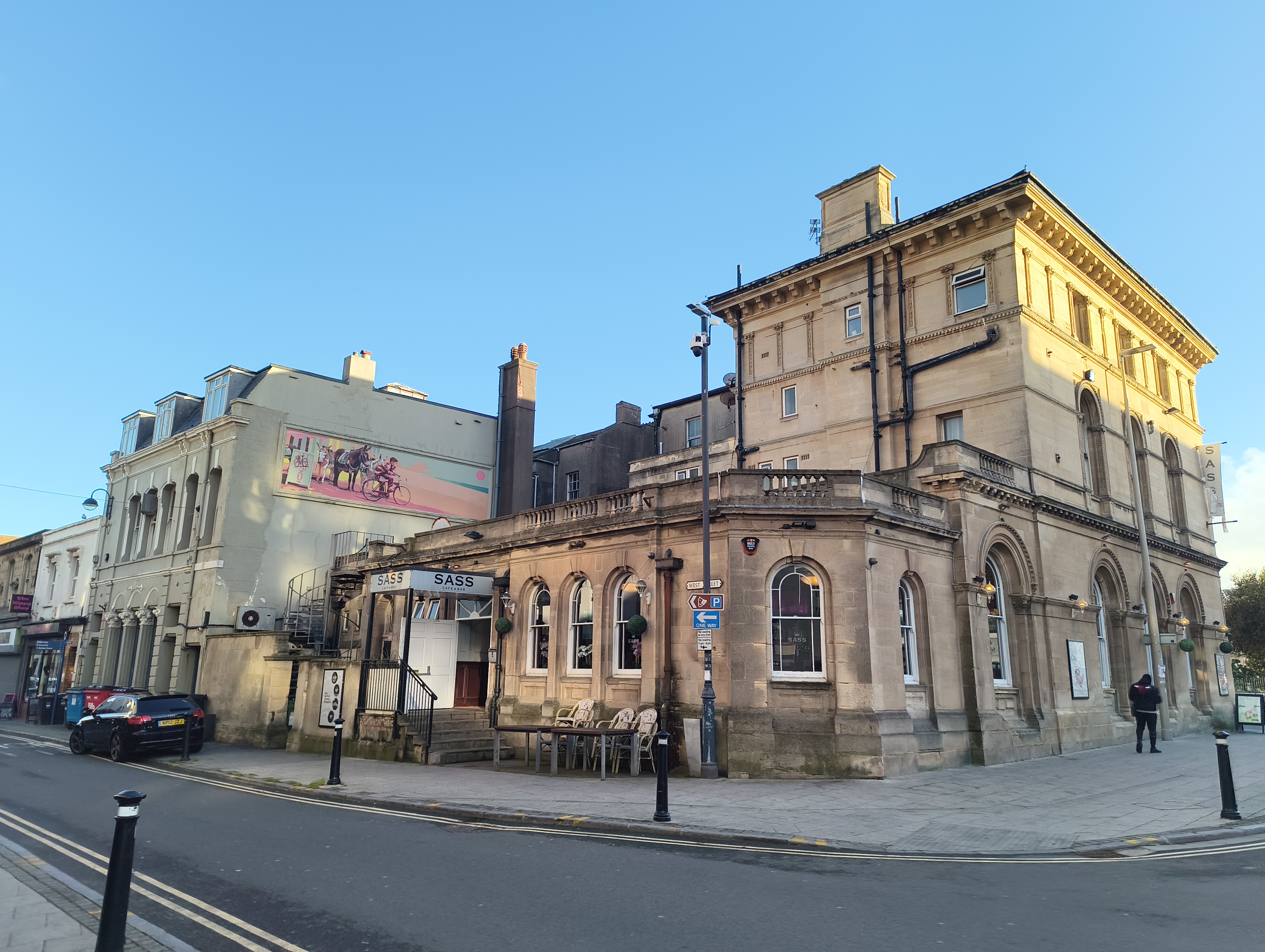 A large, historic, light-colored stone building with arched windows and a raised patio sits on a street corner under a blue sky, with a plainer building next to it featuring a mural and a sign reading "SASS."