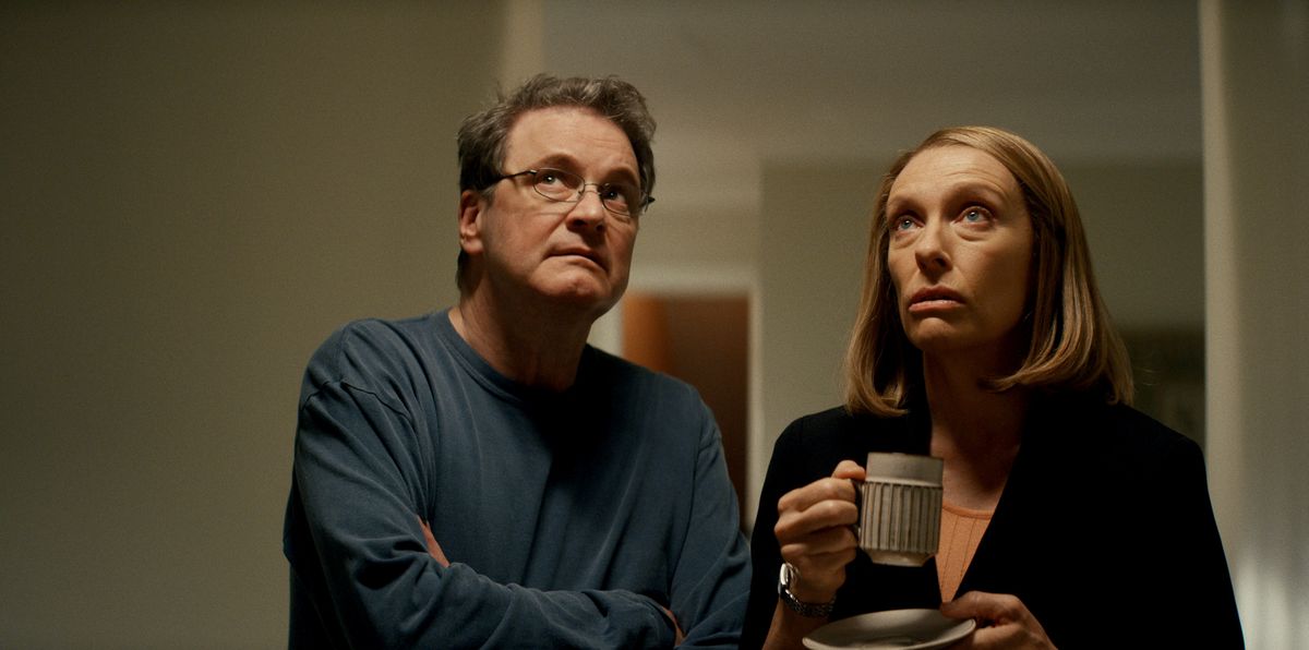 Michael Peterson (Colin Firth) and Kathleen Peterson (Toni Collette) stand in a hallway, looking up toward the ceiling. Kathleen is holding a mug in her right hand and a sauce in her left. Both of them look concerned.