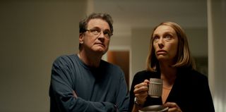 Michael Peterson (Colin Firth) and Kathleen Peterson (Toni Collette) stand in a hallway, looking up toward the ceiling. Kathleen is holding a mug in her right hand and a sauce in her left. Both of them look concerned.