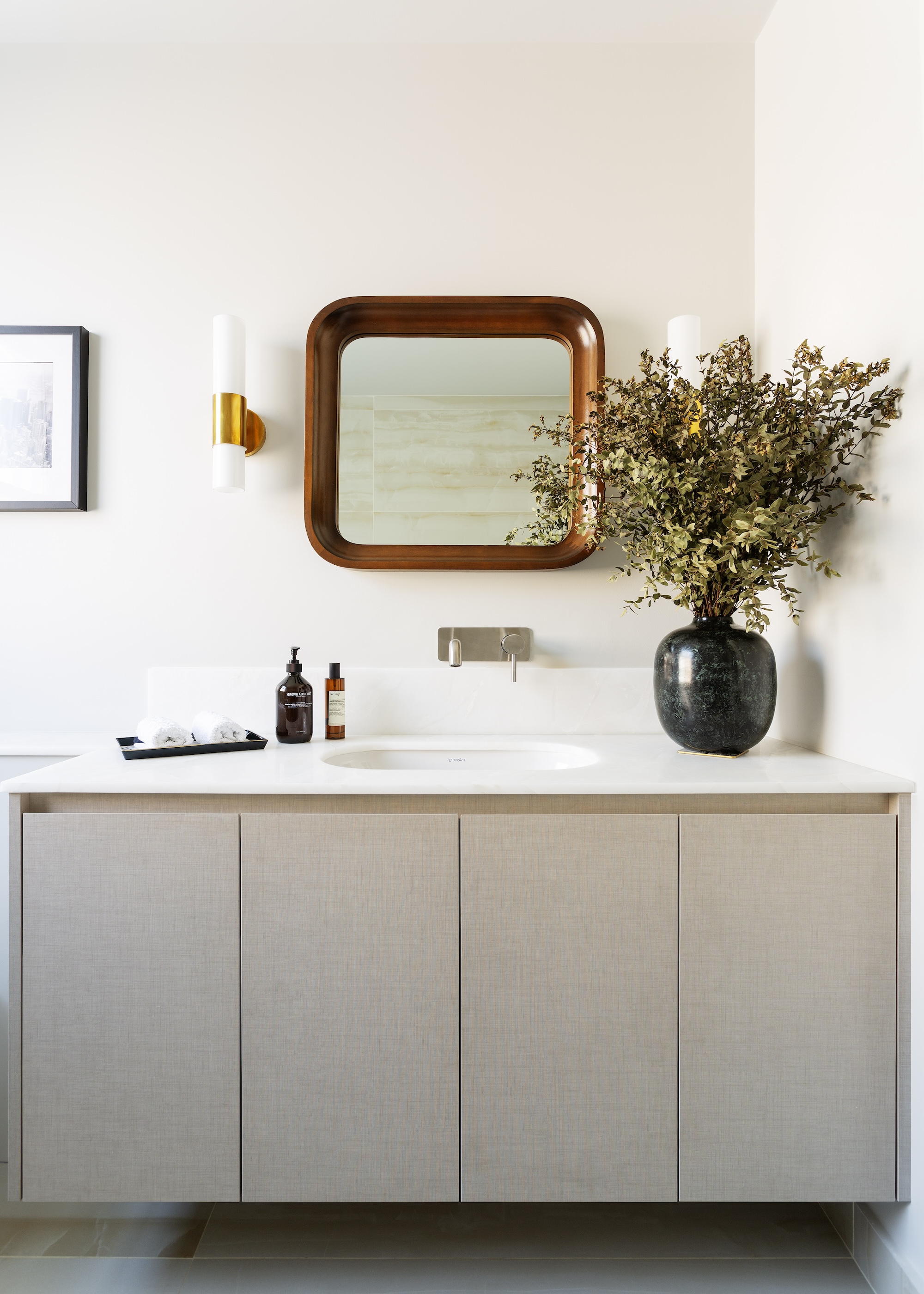 A bathroom with a vanity and a mirror above the sink as well as bathroom accessories and a black vase with plants inside of it