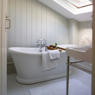 coastal bathroom with pale grey tongue and groove, large freestanding bath, white floor tiles, white bath mat, Velux window, basin