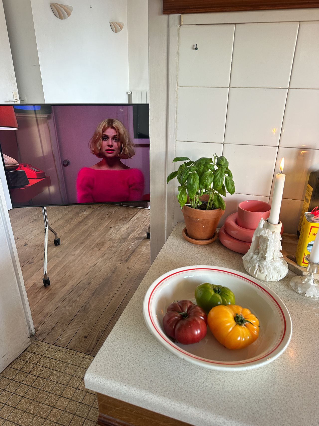 A TV on a rolling TV stand in the doorway of a white kitchen. There is a white bowl with red trim on the counter that has tomatoes in it.