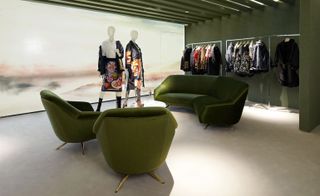 Two green chairs, a green sofa, a round glass coffee table, two mannequins and a clothing rails with clothing hanging on them.