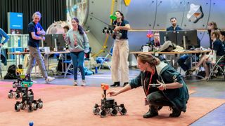 tiny six-wheel rovers roll around on an orange mat on a concrete floor. A woman in the foreground on the right kneels next to one, her hand reaching toward it. In the background another woman stands holding a rover, while others look on or sit at computer monitors.