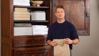 A picture of Nate Berkus, a white man with light brown short hair wearing a navy blue jumper next to a tall dark brown wooden cabinet with folded white and beige linens on the three shelves