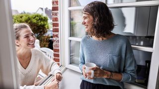 Two women laughing together looking out of garden door, holding mugs of tea