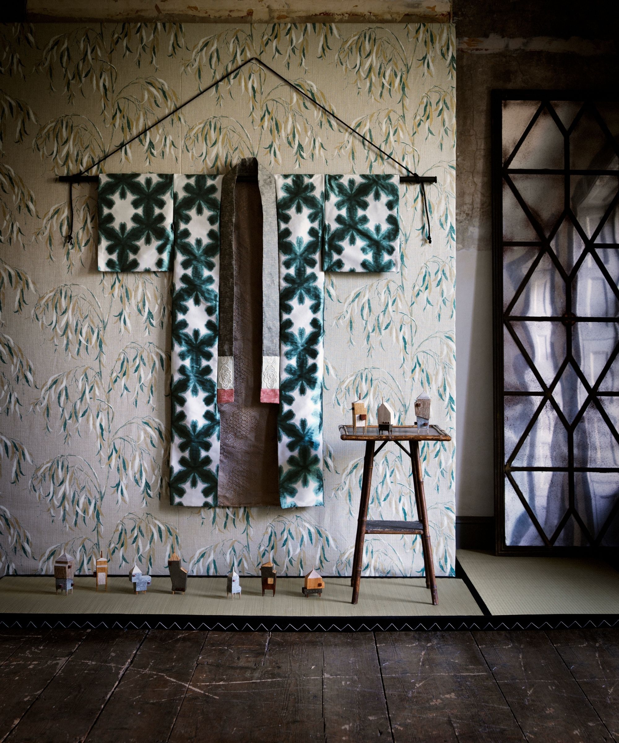 A kimono wall hanging on a floral wallpapered wall, with tatami mats, a wooden stool, and an open glass door to the right.