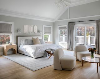 Neutral bedroom with double bed and area rug, nightstand, coffee table and two upholstered chairs, floor length drapes and view to exterior