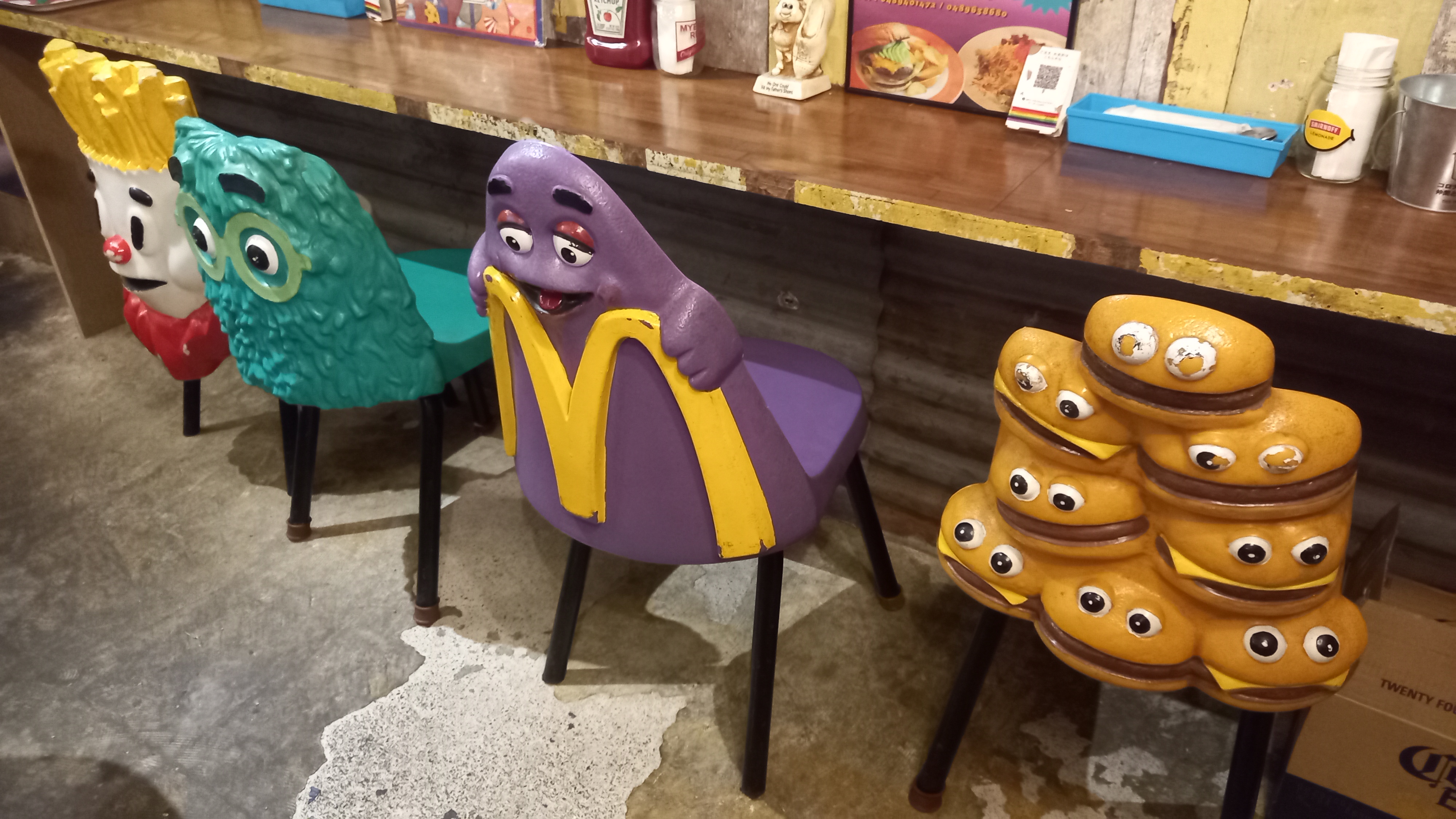 A row of original McDonaldland character chairs, featuring Grimace, the Professor and stacked burger figures, lines a wooden counter top crowded with vintage fast food packaging and condiments.