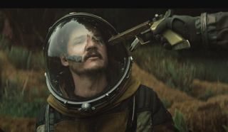 An image of Pedro Pascal as Ezra in Prospect, with someone pointing a gun to his helmeted-head