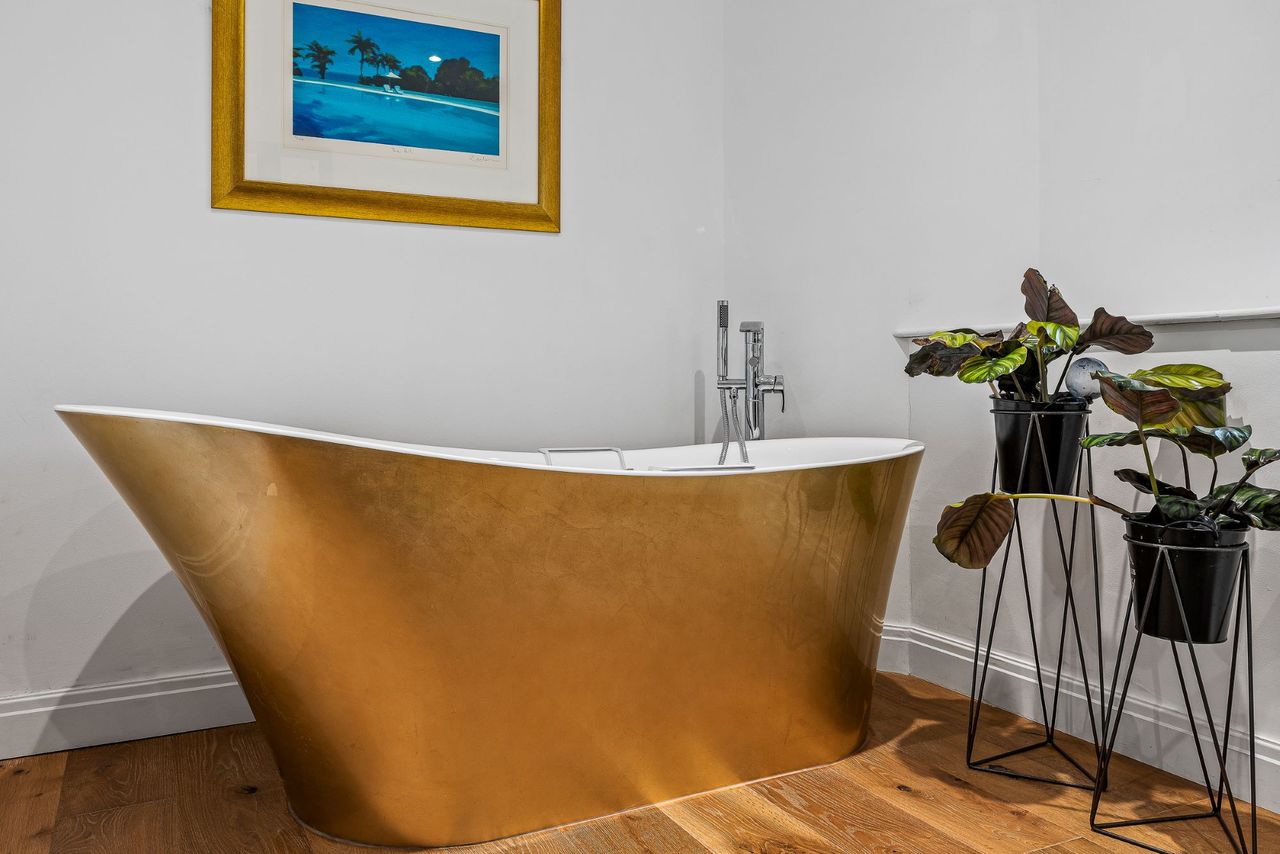 The bath is a golden exterior with white inside which is next to a potted plant and painting on the wall