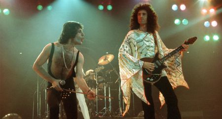 Freddie Mercury and Brian May onstage in the early 1970s; May is wearing a white patterned cape and plays his Red Special electric guitar while Mercury strikes a pose with his mic stand.