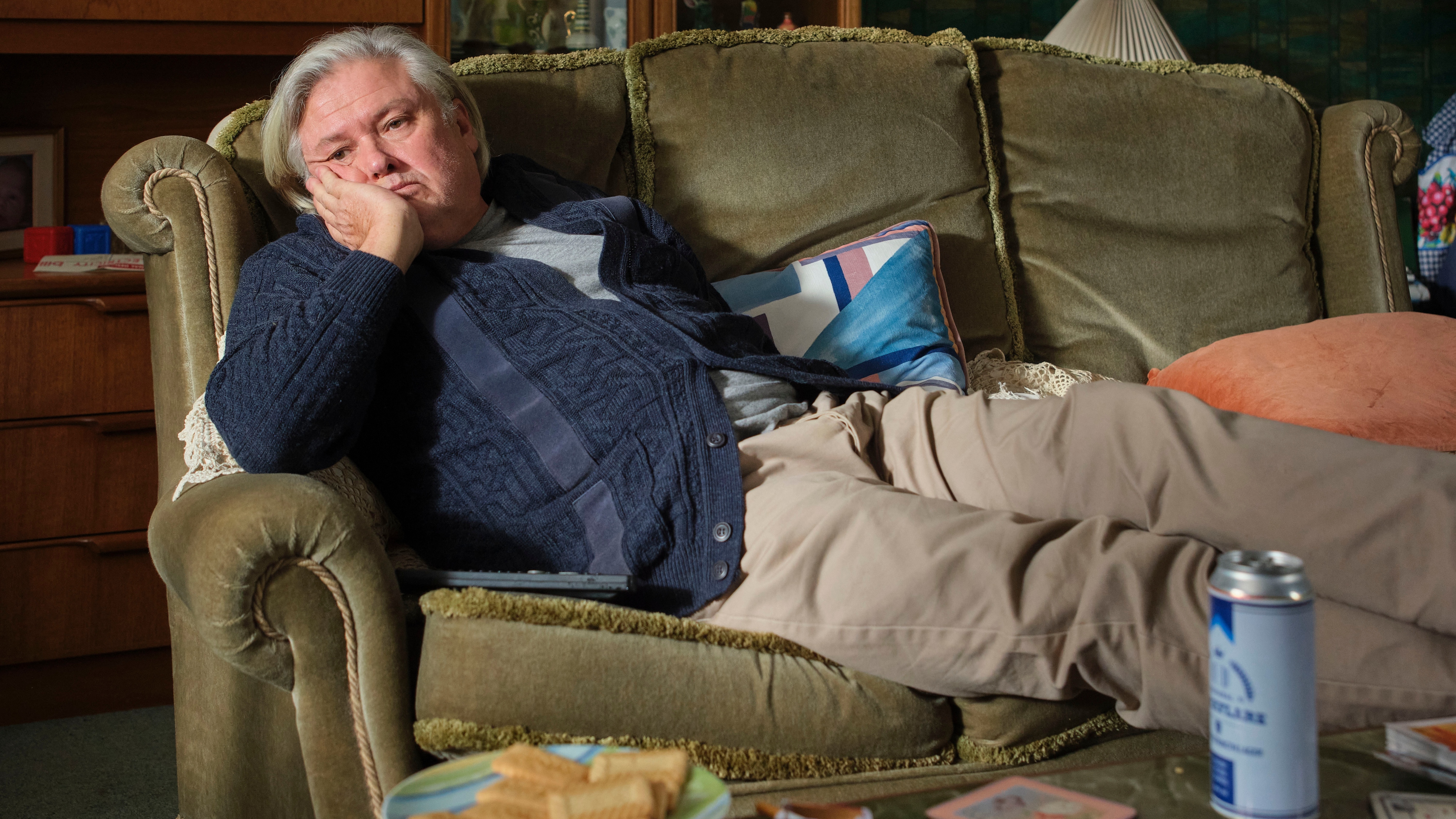Martin Parker (Conleth Hill) is lying sideways on an old green sofa with his feet up on the coffee table in front of him, leaning his chin on his hand and looking miserable. He's wearing a cardigan and chinos, and there is an open can of lager on the coffee table.