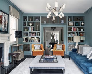 A blue living room with a wall of open shelving and orange velvet armchairs