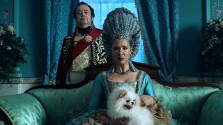 Queen Charlotte (Golda Rosheuvel) in full costume with her dog on her lap, with Brimsley (Hugh Sachs) stood behind her in Queen Charlotte: A Bridgerton story