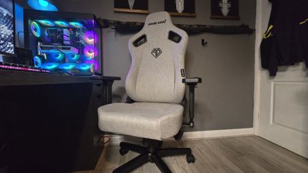 A grey gaming chair beside an RGB lighting gaming PC, desk and in an office background