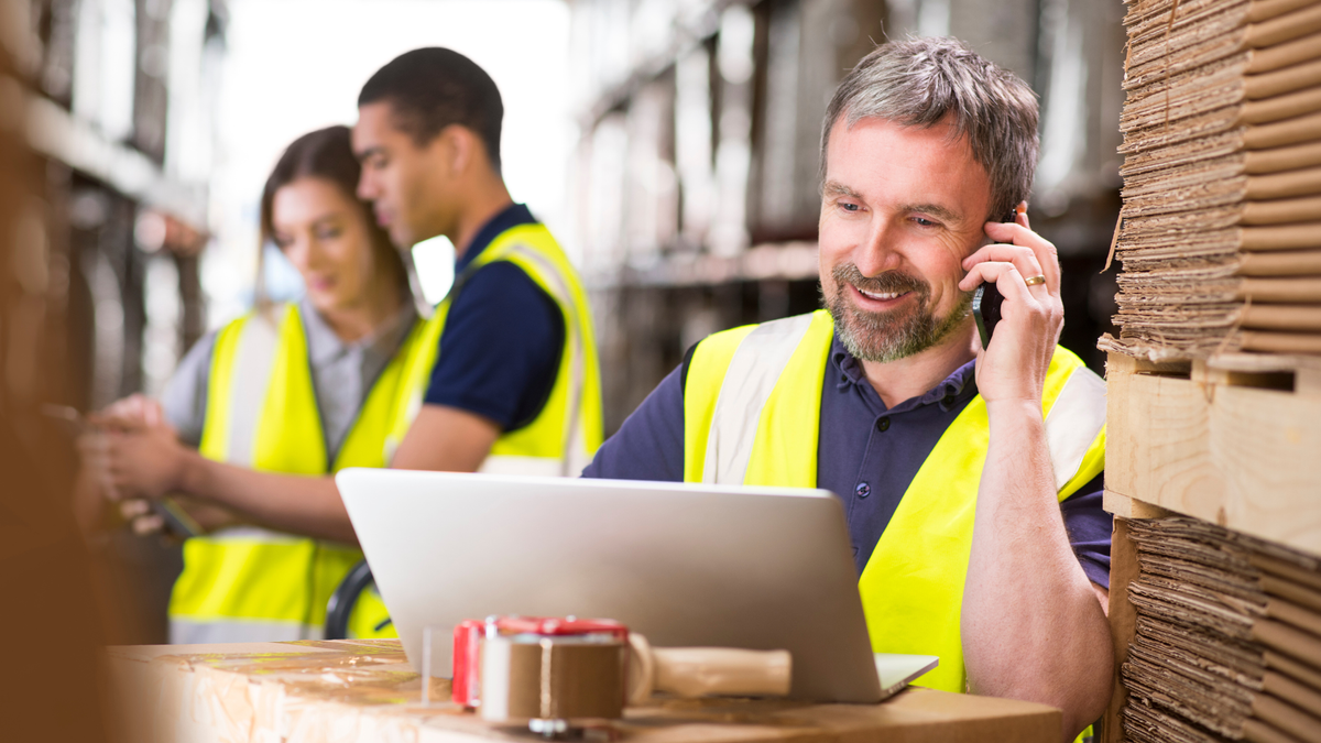 Three people wearing high vis vests are working in a warehouse. One in the foreground is on the phone and using his laptop. He looks happy.