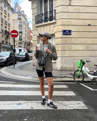 French female fashion influencer Lena Farouil poses on a Paris crosswalk wearing a gray baseball cap, black sunglasses, a houndstooth blazer, gray T-shirt, black crossbody mini bag, black biker shorts, white crewneck socks, and black Samba sneakers