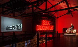 Man on a bicycle in front of a red neon sign reading "SEEK DISCOMFORT"