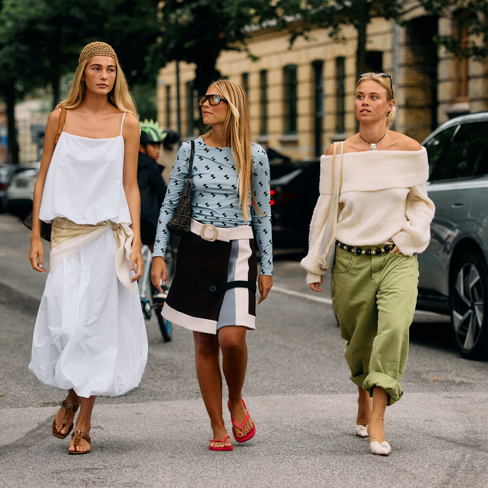 Woman walk and pose in the street. One wears a white cotton skirt set. Another wears long sleeve plue top with wrap skirt. Last one wears green utility pant and off the shoulder sweater. 