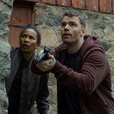 Fola Evans-Akingbola as chelsea and Gabriel Basso as peter sutherland pointing a gun and hiding behind a brick wall while investigating in the night agent season 3