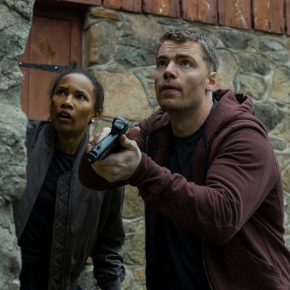 Fola Evans-Akingbola as chelsea and Gabriel Basso as peter sutherland pointing a gun and hiding behind a brick wall while investigating in the night agent season 3