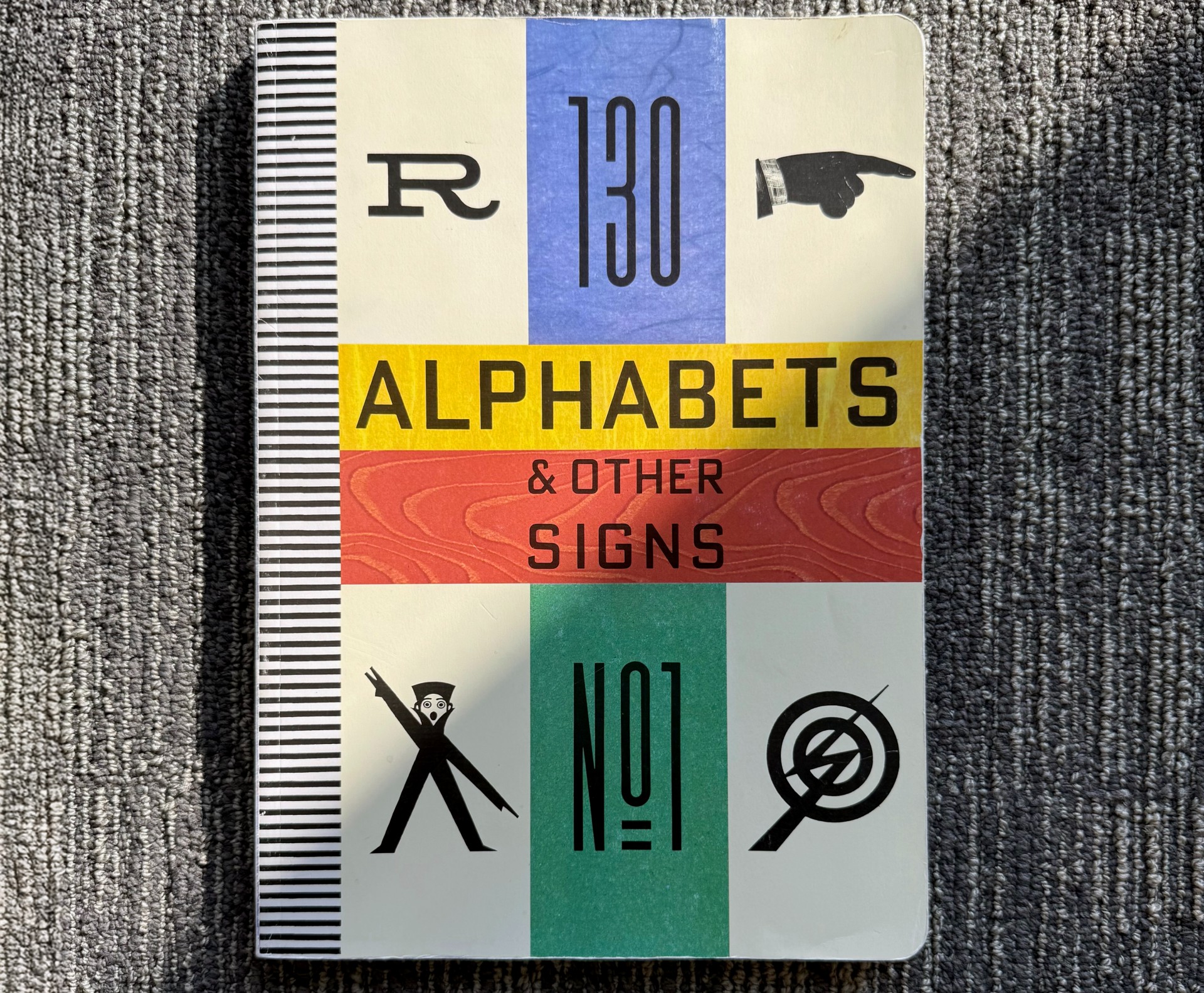 A book titled &quot;130 Alphabets &amp;amp; Other Signs&quot; with a black, blue, yellow, red, and green striped cover featuring various graphic signs and letters.