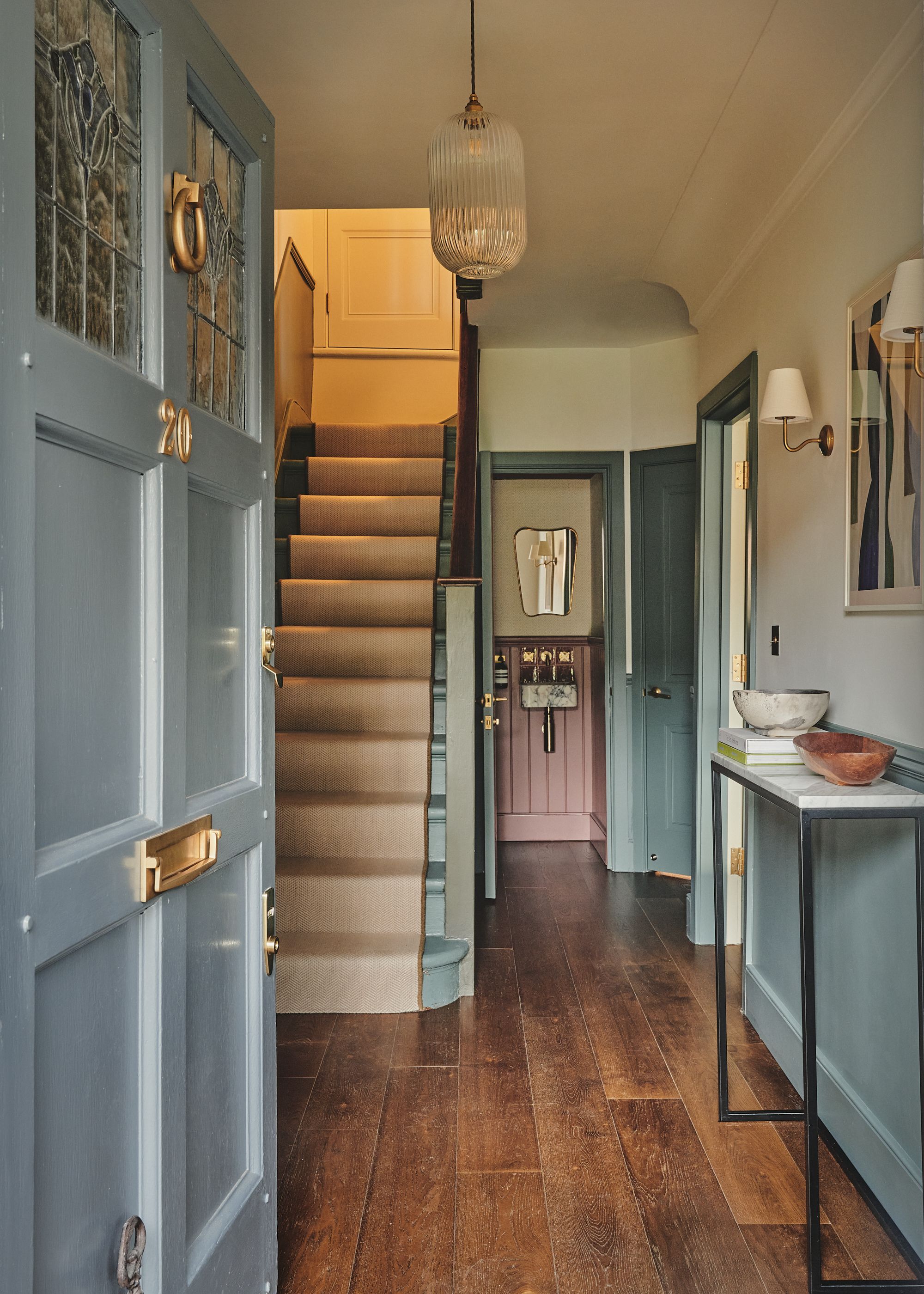 An entryway with a blue door, a staircase, wood floors, a wall lamp, and a tall side table
