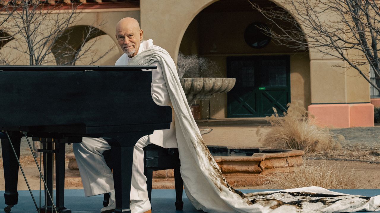 john malkovich as moretti wearing a white cape sitting at a piano outside in a still from the movie opus