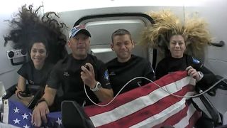 four astronauts behind a flag floating in a small spacecraft