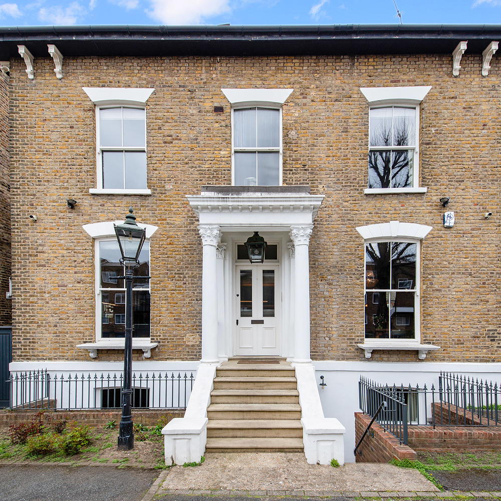 a view of the front exterior of a large brick home in London, with 5 windows and the entrance door surrounded by white column porch, with stairs leading down to the path and a traditional black lamppost at the base