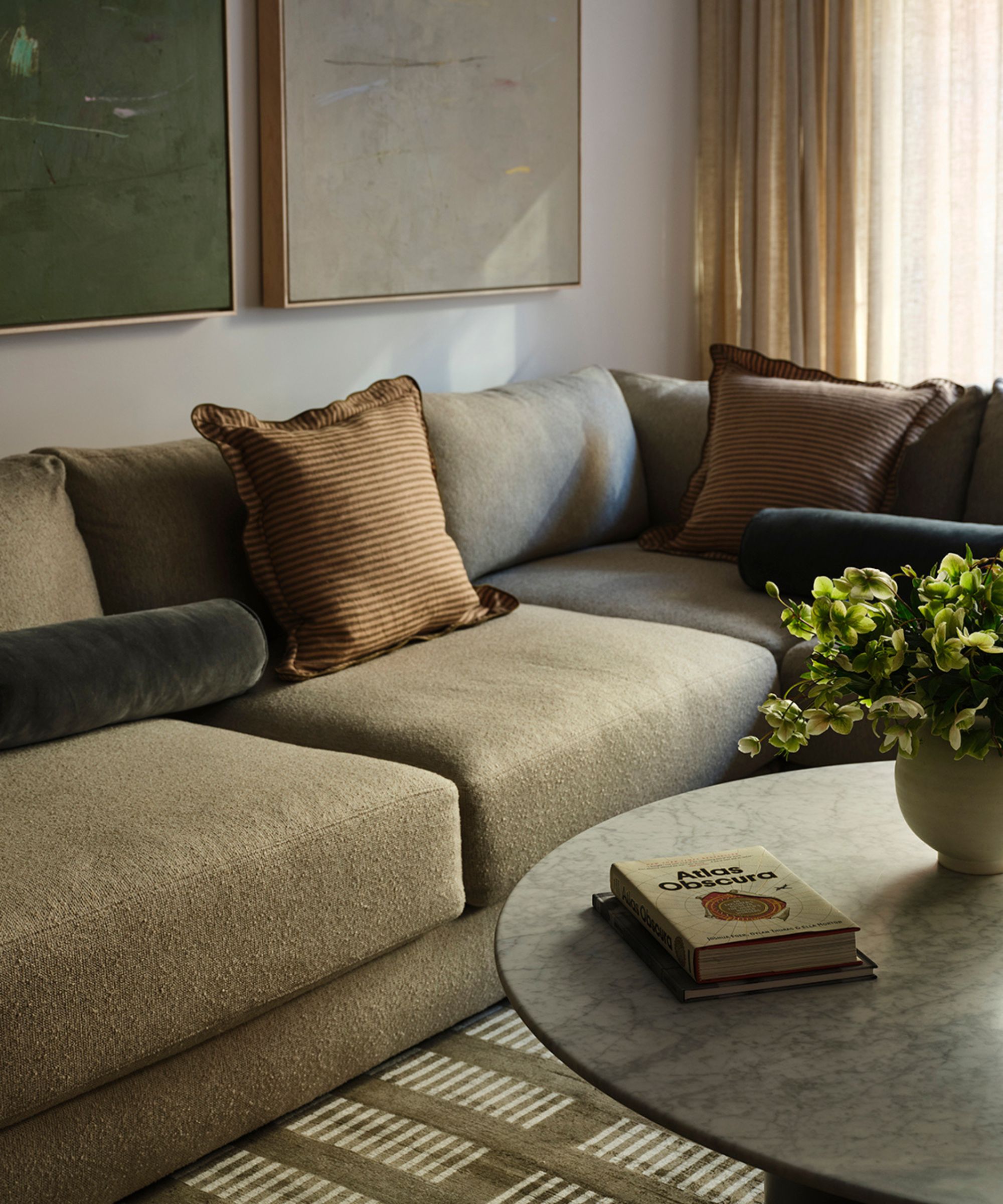 Living room with brown boucle sectional sofa, green geometric rug, a round marble coffee table with books and a plant on top, and two large paintings above the sofa in green and beige