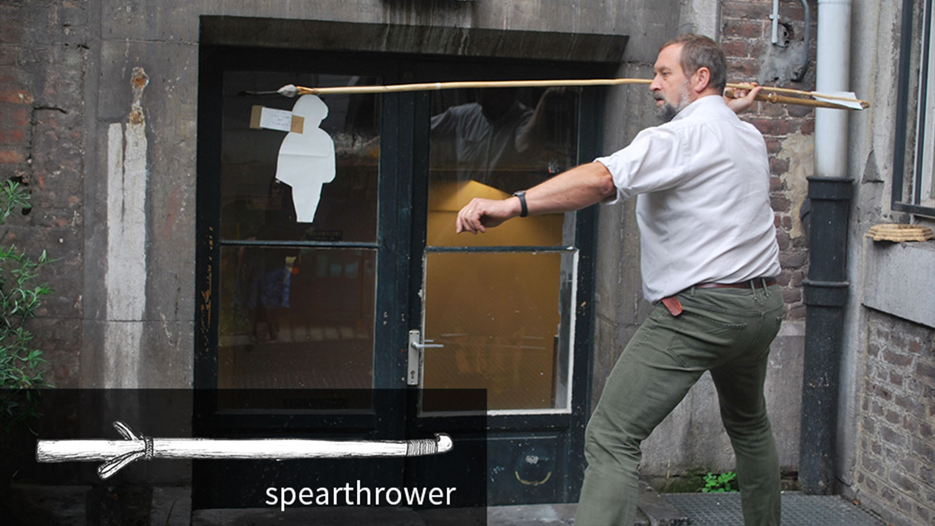 A man in a white shirt and green pants stands with a large spear about to throw it with a brick wall background.