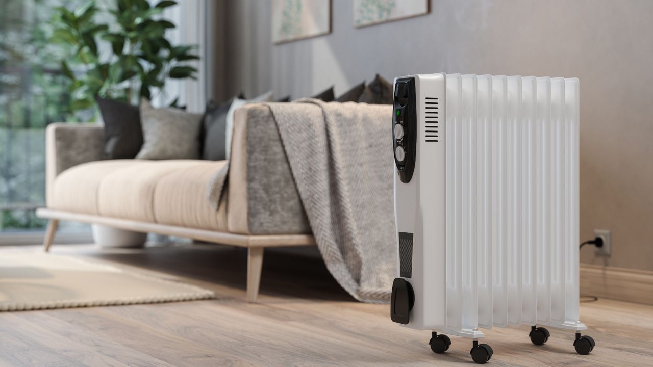 Close-up view of an electric white space heater warming a living room, with blurred cream couch and gray pillows in the background