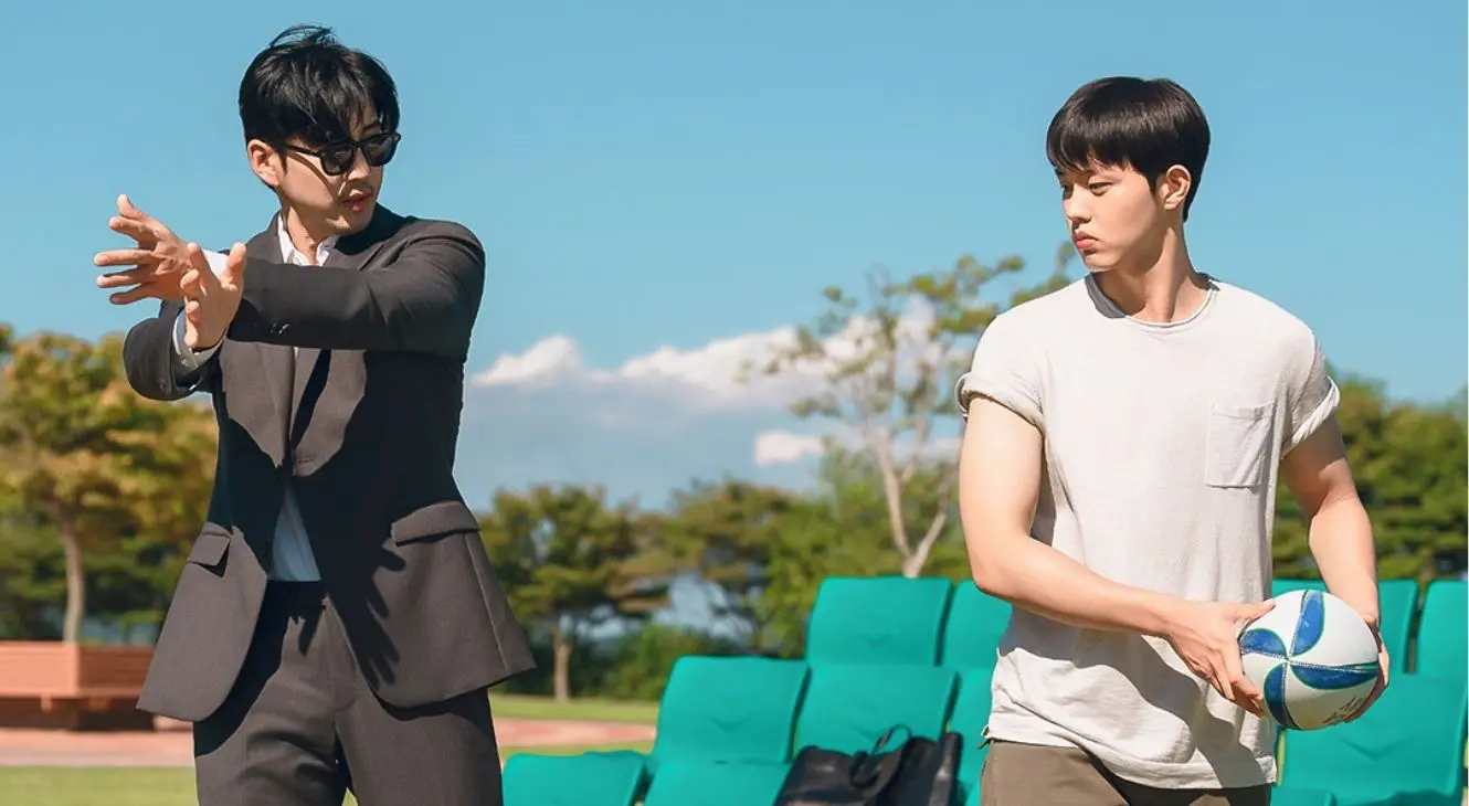 A rugby coach wearing a suit teaches a movement to a young player as they stand in front of the bleachers, in the Kdrama &#039;The Winning Try.&#039;